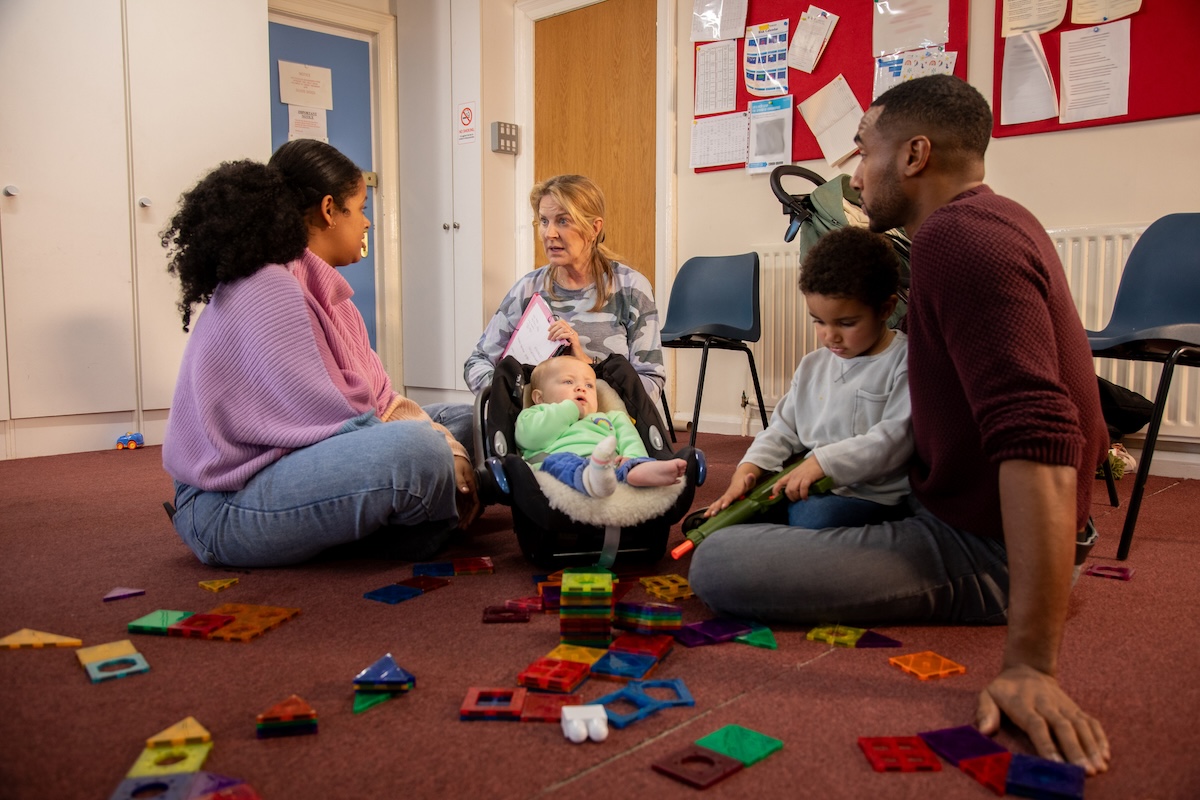 Three adults sit with two children while one plays with some toys